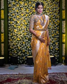 a woman standing in front of a flower wall wearing a yellow sari and gold jewelry