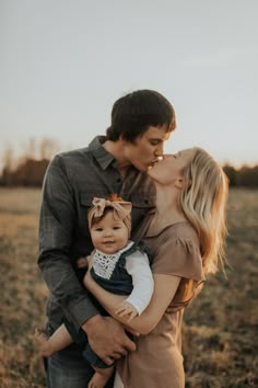 a man and woman kissing while holding a baby in an open field at sunset with the sun shining on them