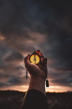 a hand holding a small clock in front of a cloudy sky at sunset or dawn