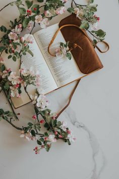 an open book on top of a marble table next to flowers and a leather bag