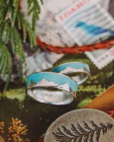 A matching wedding ring set from Staghead Designs sits on a mountain post card surrounded by props like juniper branch, leather bolo tie with pine tree, and an Idaho stamp with a bluebird. These silver wedding bands feature solid metal mountain ranges and crushed turquoise inlays sealed by a resin. Mountain Wedding Ring, Mountain Rings, Wedding Rings For Couples, Turquoise Wedding Rings, Mountain Ring, Rings For Couples, Dream Wedding Ring