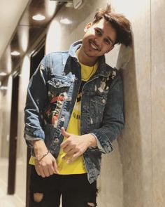 a young man leaning against a wall wearing a jean jacket and yellow t - shirt