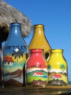 three painted jars sitting on top of a wooden table next to the ocean and thatched hut