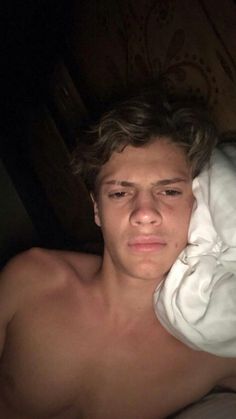 a shirtless young man laying in bed under a white blanket and looking at the camera