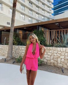 a woman in a pink romper and matching heels posing for the camera with a palm tree behind her