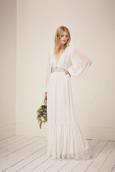 a woman in a long white dress holding a bouquet and posing for the camera with her hands on her hips