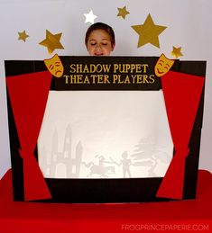 a young boy is holding up a shadow puppet theatre sign with stars above him and behind it, there is a red table cloth