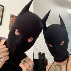 two people wearing black masks with horns and piercings on their heads, one is holding a ring in front of the other's face