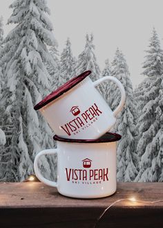 two white coffee mugs sitting on top of a wooden table in front of snow covered trees