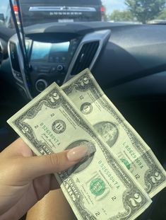 a person holding up two bills in their hand next to the dashboard of a car