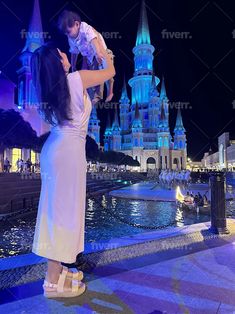 a woman holding a baby in front of a castle at night with the lights on