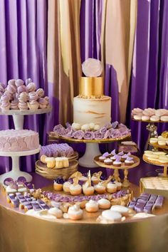 a table topped with lots of different types of cakes and cupcakes next to purple curtains