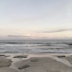 an empty beach with waves coming in from the ocean and rocks on the sand, under a cloudy sky