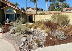 a house with lots of plants and rocks in front of the house on a sunny day