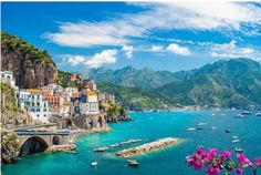 boats are floating in the blue water near some buildings and mountains with green hills behind them