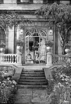 an old black and white photo of a building with flowers on the front porch, and stairs leading up to it