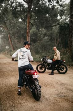 two men riding motorcycles on a dirt road in the woods with one man sitting on his motorcycle