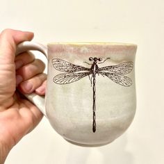 a hand holding a coffee cup with a dragonfly on it's outside side