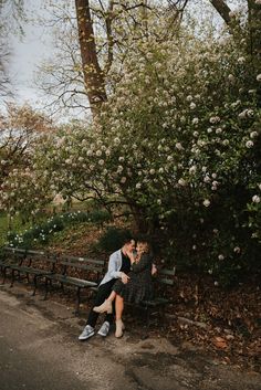 two people sitting on a bench in front of some trees and bushes with white flowers