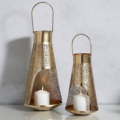 two gold metal candle holders with white candles in them sitting on a table next to each other