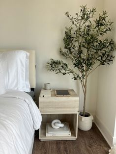 a bed with white sheets and pillows next to a small table with a plant on it