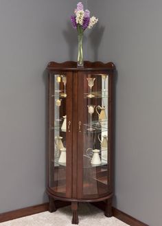 a corner display cabinet with glass doors and shelves on the top, in front of a gray wall