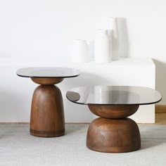 two wooden tables with glass tops in front of a white wall and vases on the floor