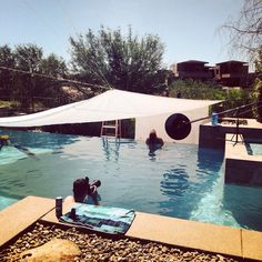 two people are swimming in an outdoor pool under a white tarp and some trees