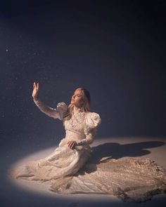 a woman in a white dress sitting on the ground with her hand up to the sky