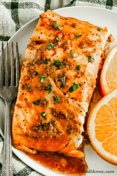 a white plate topped with fish covered in sauce next to sliced oranges and a fork