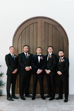 a group of men standing next to each other in front of a wooden door wearing tuxedos