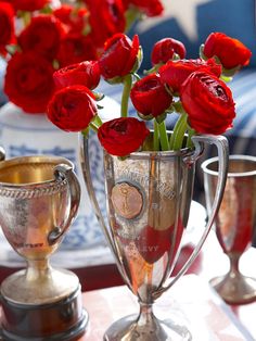 red roses are in a silver vase on a table with two cups and one is empty