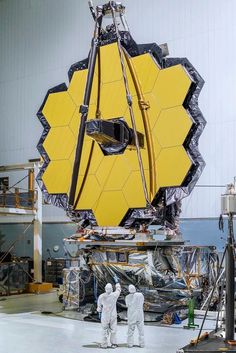 two men in white suits standing next to a large yellow object on top of a metal structure