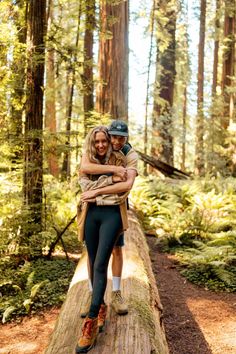 a man and woman are hugging on a log in the middle of a forest with tall trees