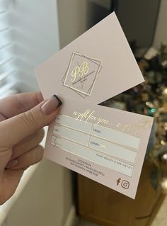 a person holding up two pink and gold business cards in front of a christmas tree