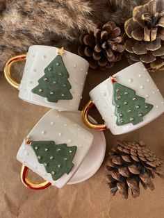 three mugs with christmas trees on them sitting next to a pine cone and cones