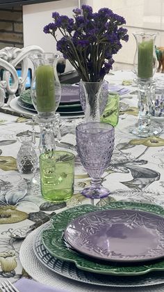 the table is set with purple flowers and green glassware, plates and silverware