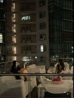 two women sitting on white couches in front of a tall building at night time