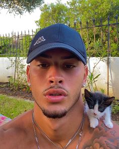 a shirtless man holding a small kitten on his shoulder and wearing a baseball cap