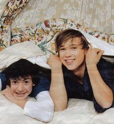 two young men laying on top of each other in bed under covers and pillows,