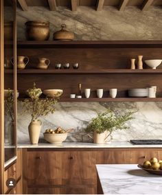 a kitchen with marble counter tops and wooden shelves filled with bowls, cups, vases and fruit