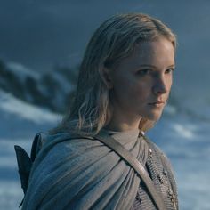 a woman with long blonde hair wearing a cape and standing in front of snow covered mountains