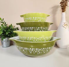 four green glass bowls with white flowers on them sitting next to a vase and potted plant