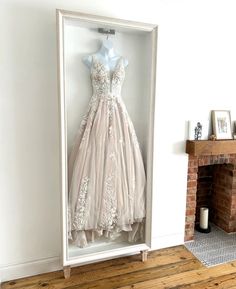 a wedding dress in a white frame on a wooden floor next to a fire place