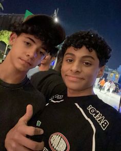 two young men standing next to each other in front of a building at night with their thumbs up