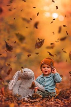 a little boy sitting in the leaves with a stuffed animal next to him and falling leaves all around him