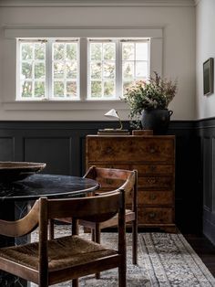 a dining room table with chairs and a vase on top of it in front of two windows