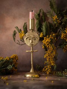 a gold candle holder with a face on it and yellow flowers in the back ground