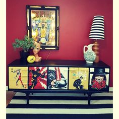 a sideboard with pictures on it in front of a red wall and a lamp