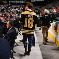 a hockey player is walking down the court with his jersey draped over his shoulders, and people are sitting in chairs behind him